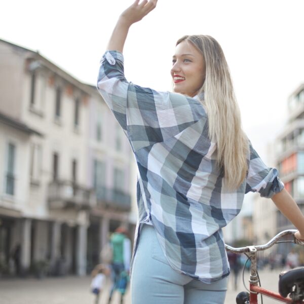 Back View Photo of Smiling Woman in Checkered Shirt and Blue Denim Jeans Standing with a Bicycle Waving