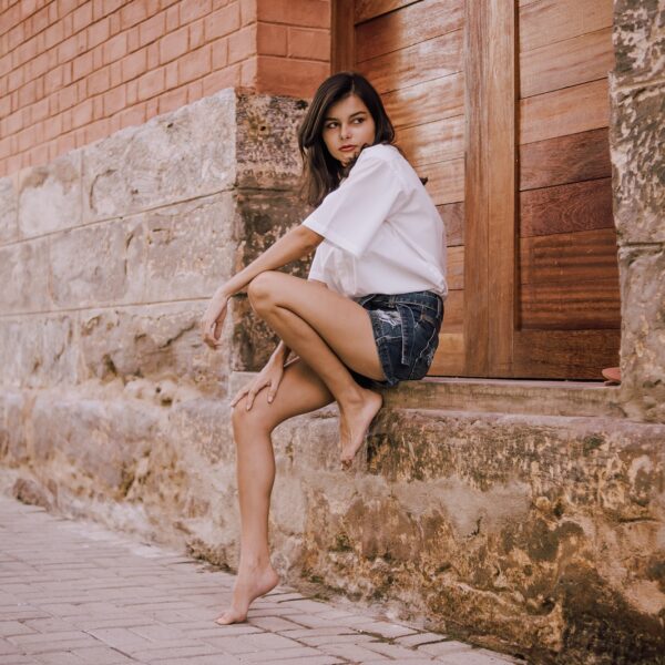 Side view of distrustful young barefoot ethnic woman in stylish cloths sitting on weathered surface of old stone building near pavement and looking away