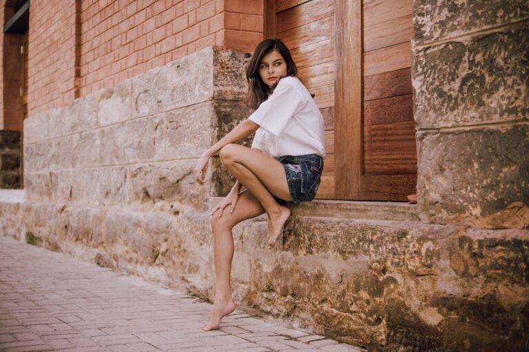 Side view of distrustful young barefoot ethnic woman in stylish cloths sitting on weathered surface of old stone building near pavement and looking away