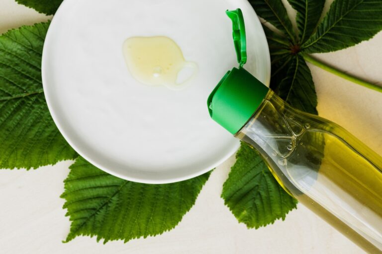 Top view of open bottle of cosmetic oil placed flat on table near white plate with small amount of oil on it on big fresh green leaf