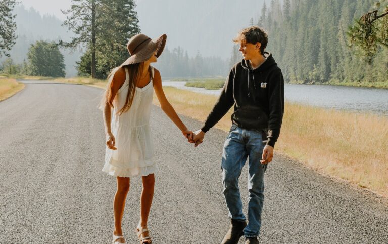 Anonymous female teenager in stylish sundress and hat holding trendy smiling boyfriend by hand while looking at each other on roadway