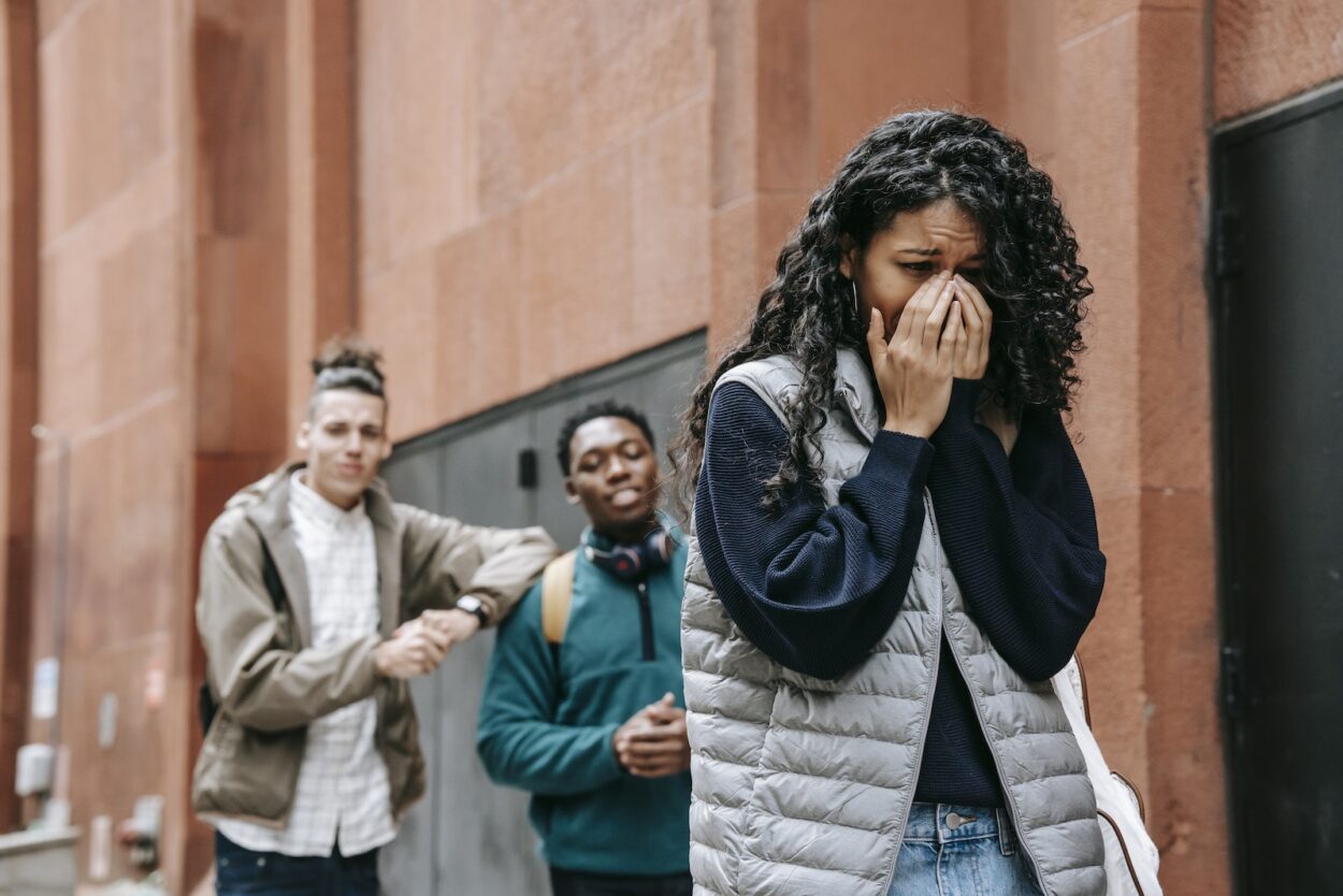 From below of despaired young ethnic female student covering mouth with hands while crying on street after being bullied by multiracial classmates