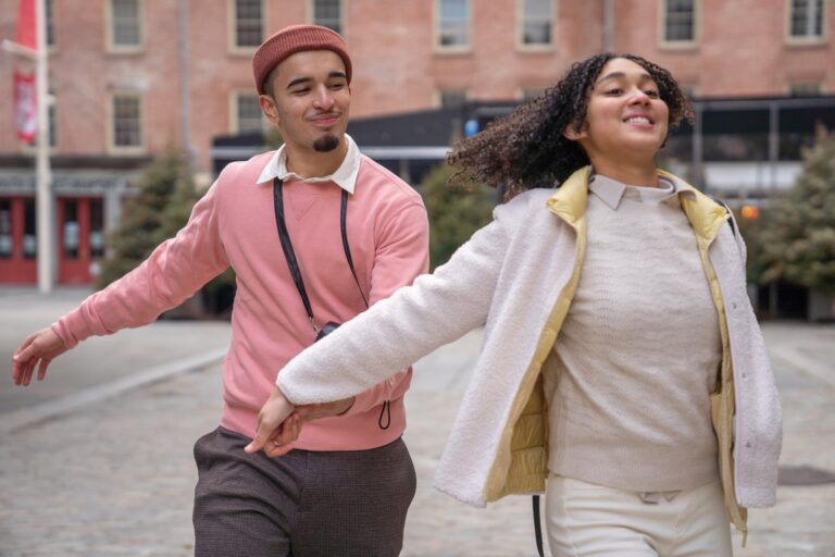 Cheerful young Hispanic couple in stylish clothes holding hands and running on city square during romantic date